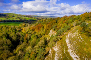 A vibrant autumn landscape at Loggerheads, North East Wales, featuring a wooded hillside with green, yellow, and orange foliage. A rocky cliff edge contrasts with the dense forest below, while rolling green fields and distant hills stretch across the background under a partly cloudy sky. A few hikers are seen exploring the trail along the ridge.