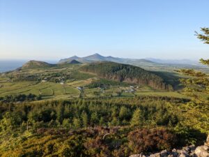 Scenic view of the Llyn Peninsula, featuring rolling green fields, clusters of trees, and distant mountains under a clear blue sky. The landscape is dotted with farm buildings and winding country roads, with the coastline visible on the left.